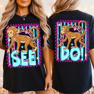 May include: Black t-shirt featuring a cartoon monkey illustration. The front of the shirt has the word "SEE" in blue bubble letters, and the back has the word "DO!" in the same style. The monkey is brown with a pink, blue, and yellow border.
