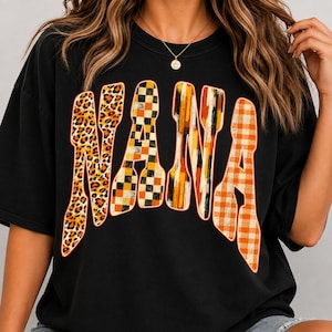 May include: A black short-sleeved t-shirt featuring the word NANA printed in large, stylized letters across the chest. Each letter has a distinct pattern: leopard print, a black and orange checkerboard, vertical orange brushstrokes, and orange gingham.