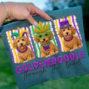 May include: Teal fabric featuring three embroidered Goldendoodles in Mardi Gras attire. Each dog wears a different festive accessory: a jester hat, a mask, and a top hat. The text "GOLDENDOODLE Through The beads" is embroidered below the dogs.