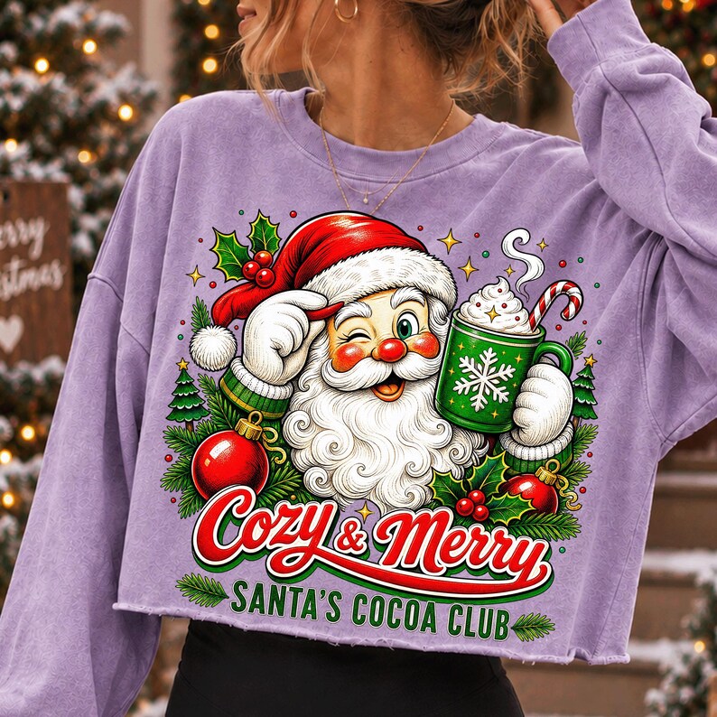 K&ouml;nnte beinhalten: Ein hellviolettes Langarm-Sweatshirt mit einer Grafik von einem zwinkernden Weihnachtsmann, der eine gr&uuml;ne Tasse mit wei&szlig;em Schneeflocken-Design h&auml;lt. Der Text lautet Cozy & Merry SANTA'S COCOA CLUB, umgeben von Stechpalmen, Tannen und roten Kugeln.