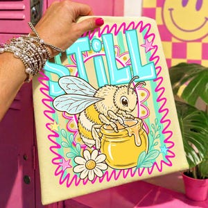 May include: A light yellow towel with a cartoon bee design. The bee is perched on a honey pot, with the word "STILL" in blue letters above. The design includes flowers, stars, and a pink wavy border.
