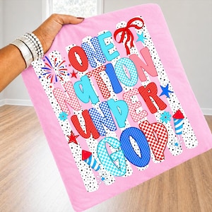 Puede incluir: Manta rosa con el texto "ONE NATION UNDER GOD" en letras rojas, azules y rosas. La manta tiene un borde punteado y presenta dise&ntilde;os de fuegos artificiales y estrellas. La manta est&aacute; sujeta por una persona.
