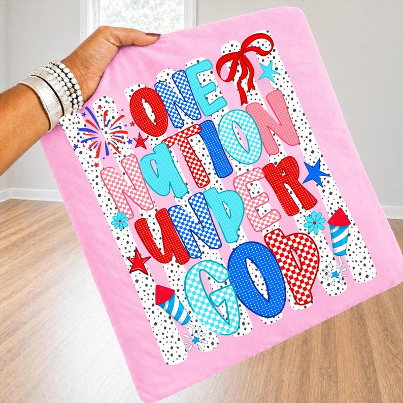 Puede incluir: Manta rosa con el texto "ONE NATION UNDER GOD" en letras rojas, azules y rosas. La manta tiene un borde punteado y presenta dise&ntilde;os de fuegos artificiales y estrellas. La manta est&aacute; sujeta por una persona.