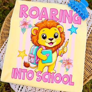 May include: A light yellow t-shirt featuring a cartoon lion with a brown mane, holding a yellow pencil and a teal book. The lion wears a pink backpack. The text reads ROARING INTO SCHOOL in pink letters, surrounded by pink flowers and blue and yellow stars.