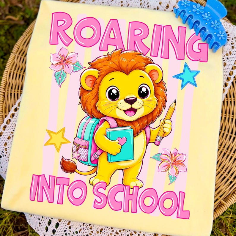 May include: A light yellow t-shirt featuring a cartoon lion with a brown mane, holding a yellow pencil and a teal book. The lion wears a pink backpack. The text reads ROARING INTO SCHOOL in pink letters, surrounded by pink flowers and blue and yellow stars.