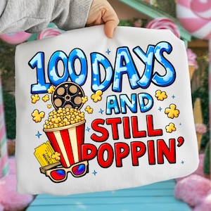Puede incluir: Camiseta blanca con un gr&aacute;fico colorido. El dise&ntilde;o presenta el texto "100 DAYS AND STILL POPPIN'" en azul y rojo, junto con palomitas de ma&iacute;z, una bobina de pel&iacute;cula, gafas 3D y una entrada de cine.