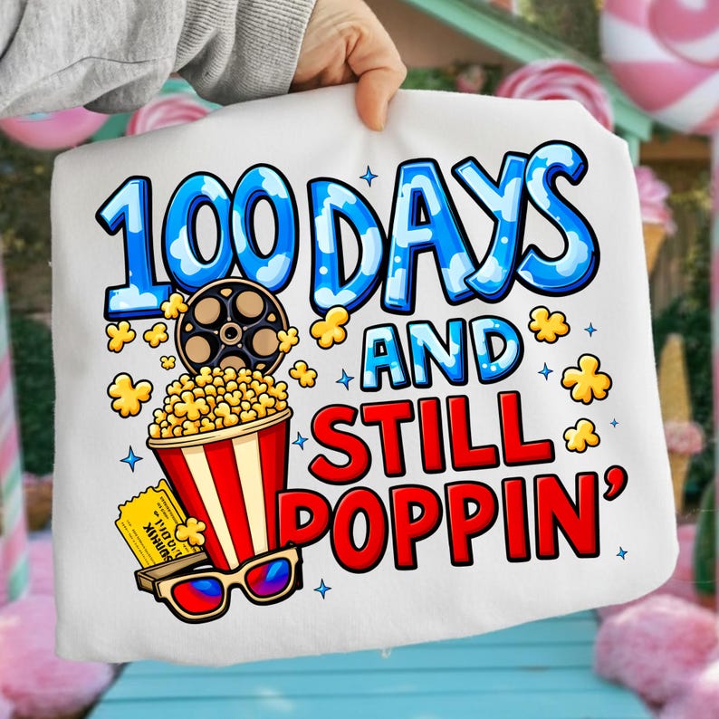 Puede incluir: Camiseta blanca con un gr&aacute;fico colorido. El dise&ntilde;o presenta el texto "100 DAYS AND STILL POPPIN'" en azul y rojo, junto con palomitas de ma&iacute;z, una bobina de pel&iacute;cula, gafas 3D y una entrada de cine.