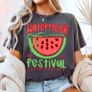 以下が含まれることがあります： スイカのデザインが施された濃いグレーのTシャツ。「Watermelon Summer Festival」の文字が赤と緑で描かれ、スイカのスライスのイラストと「Sweet and Fresh」の文字がデザインされています。