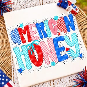 May include: A white t-shirt featuring the text AMERICAN HONEY in a multi-colored, patterned font. The letters are decorated with red, blue, and pink polka dots and checkerboard patterns, accented with small stars, flowers, and a firework graphic.