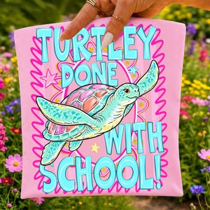 以下が含まれることがあります： ピンク色のTシャツ。水色、ピンク、オレンジ色のカメのイラストが描かれています。「TURTLEY DONE WITH SCHOOL!」の文字はターコイズ色。星と波状のピンク色の縁取りのデザインです。