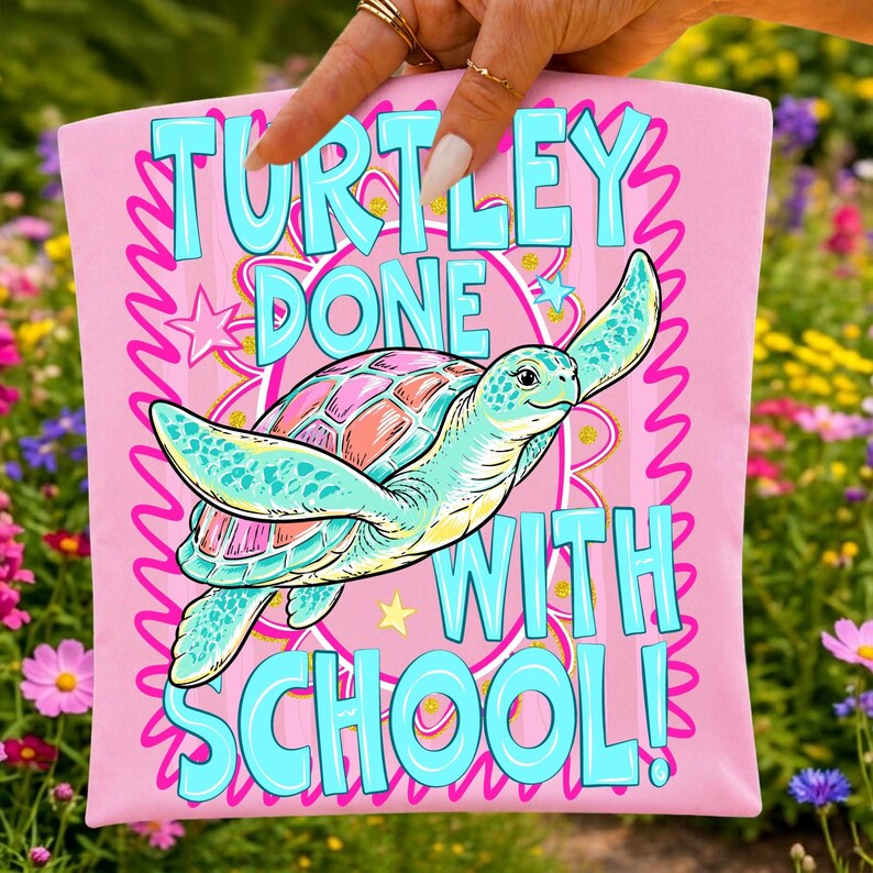以下が含まれることがあります： ピンク色のTシャツ。水色、ピンク、オレンジ色のカメのイラストが描かれています。「TURTLEY DONE WITH SCHOOL!」の文字はターコイズ色。星と波状のピンク色の縁取りのデザインです。
