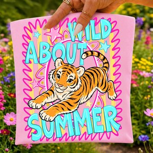 K&ouml;nnte beinhalten: Rosa Tragetasche mit einer Cartoon-Tiger-Illustration und den Worten "WILD ABOUT SUMMER" in T&uuml;rkis. Das Design umfasst Sterne und Wellenlinien in Gelb und Rosa. Der Tiger ist orange und schwarz.