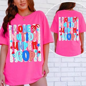 Puede incluir: Una camiseta rosa brillante con la frase "ONE NATION UNDER GOD" en letras coloridas y estampadas. El dise&ntilde;o incluye estrellas, fuegos artificiales y lazos, sobre rayas verticales blancas. La parte trasera de la camiseta refleja el dise&ntilde;o frontal.