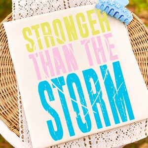May include: A cream-colored canvas with the words "STRONGER THAN THE STORM" printed in yellow, pink, and blue. A blue hair clip with flower details rests on the top right corner. The canvas is placed on a wicker surface.