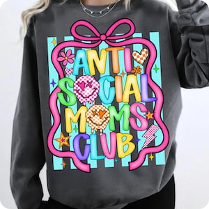 K&ouml;nnte beinhalten: Dunkelgraues Sweatshirt mit einem farbenfrohen Aufdruck "ANTI SOCIAL MOMS CLUB". Das Design umfasst ein rosa Band, Blumen, Herzen, Sterne und Smiley-Gesichter. Der Text ist in verschiedenen leuchtenden Farben gehalten.