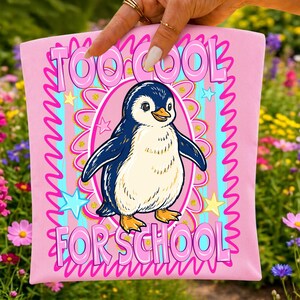 K&ouml;nnte beinhalten: Rosafarbenes T-Shirt mit einer Cartoon-Pinguin-Illustration. Der Pinguin ist blau, wei&szlig; und gelb und befindet sich in einem rosa Oval. Der Text "TOO COOL FOR SCHOOL" ist rosa, wei&szlig; umrandet und umgibt den Pinguin.