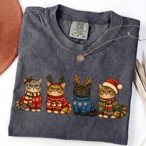 K&ouml;nnte beinhalten: Ein anthrazitfarbenes T-Shirt mit einer Grafik von vier Katzen in festlicher Kleidung. Die Katzen tragen Brillen, Pullover, Schals und Rentiergeweihe. Das T-Shirt hat ein Comfort Colors-Etikett am Kragen und eine d&uuml;nne silberne Halskette.