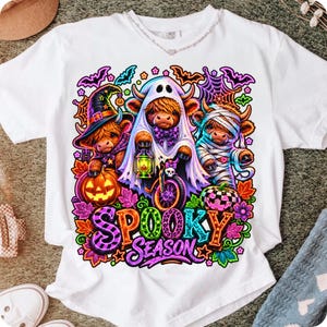 Może przedstawiać: Biały t-shirt z kr&oacute;tkim rękawem z kolorową grafiką przedstawiającą trzy krowy rasy Highland przebrane na Halloween. Jedna ma kapelusz czarownicy, druga jest duchem z latarnią, a trzecia mumią. Poniżej znajduje się napis Spooky Season.