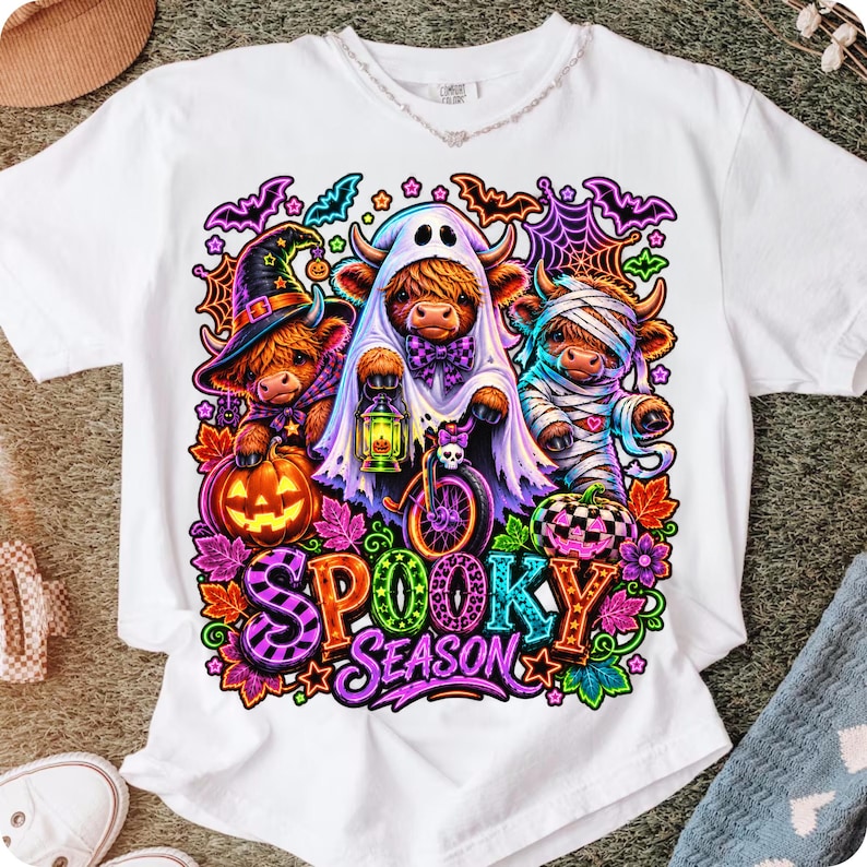 Może przedstawiać: Biały t-shirt z kr&oacute;tkim rękawem z kolorową grafiką przedstawiającą trzy krowy rasy Highland przebrane na Halloween. Jedna ma kapelusz czarownicy, druga jest duchem z latarnią, a trzecia mumią. Poniżej znajduje się napis Spooky Season.