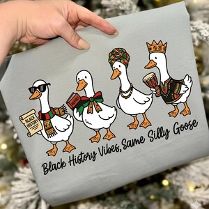 May include: A light gray fabric with a whimsical illustration of four white geese. Each goose is adorned with accessories like sunglasses, a scarf, a book, and a crown. The text "Black History Vibes, Same Silly Goose" is printed below the geese.