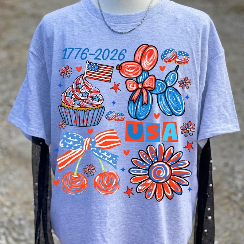 Pode incluir: T-shirt cinzento claro com um design patri&oacute;tico colorido. O design inclui um cupcake com uma bandeira, um c&atilde;o de bal&atilde;o, cerejas e uma flor, todos em vermelho, branco e azul. O texto "USA" e "1776-2026" tamb&eacute;m est&atilde;o presentes.