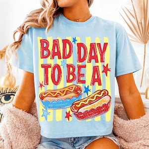 K&ouml;nnte beinhalten: Hellblaues T-Shirt mit dem Text "BAD DAY TO BE A" in roten Buchstaben. Das Design umfasst zwei Hotdogs und Sternakzente. Das Shirt hat gelbe und blaue vertikale Streifen im Hintergrund.