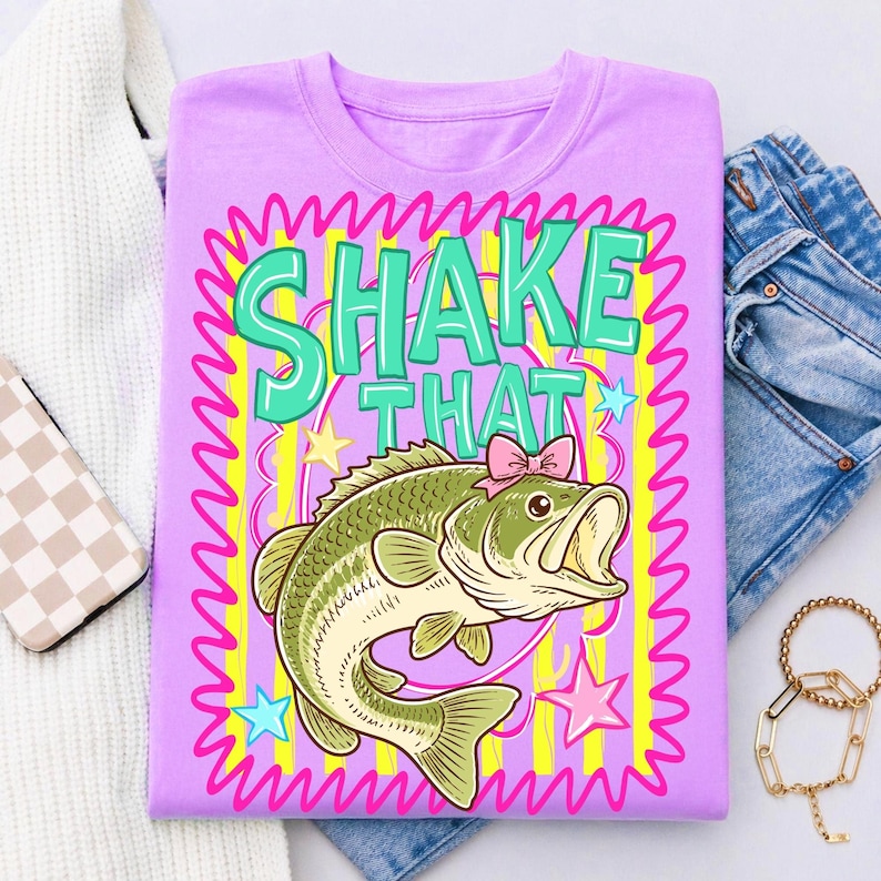 Op de afbeelding: Een lila T-shirt met een cartoon bas met een roze strik, tegen een kleurrijk ontwerp. De tekst "SHAKE THAT" is groen, omlijnd in wit en omlijst door roze zigzags en gele strepen. Blauwe en roze sterren maken het ontwerp compleet.