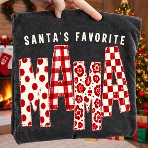 K&ouml;nnte beinhalten: Ein anthrazitfarbenes T-Shirt mit dem wei&szlig;en Schriftzug SANTA'S FAVORITE &uuml;ber dem Wort MAMA in gro&szlig;en Buchstaben. Jeder Buchstabe von MAMA zeigt ein anderes rot-wei&szlig;es Muster, darunter Punkte, Karomuster, Blumen und ein Schachbrettmuster.