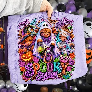 Może przedstawiać: Jasnofioletowy materiał z grafiką trzech kr&oacute;w rasy Highland przebranych na Halloween: czarownica z dynią, duch na rowerze z latarnią oraz mumia. Napis Spooky Season wykonany jest kolorowymi literami na środku nadruku.