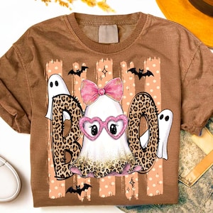 May include: A brown t-shirt featuring a graphic of a ghost wearing pink heart-shaped glasses and a pink bow. The word BOO is written in large letters with a leopard print pattern. The design includes small bats, stars, and polka dot stripes.