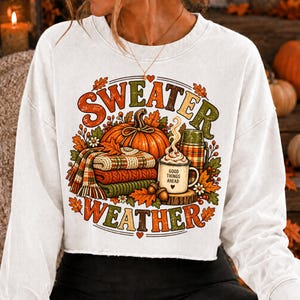 Puede incluir: Una sudadera blanca de manga larga con un estampado gr&aacute;fico colorido y el texto "SWEATER WEATHER". El dise&ntilde;o incluye una calabaza naranja grande, una pila de mantas de punto dobladas, un termo a cuadros y una taza con "GOOD THINGS AHEAD".