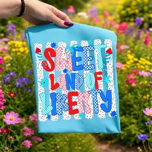 May include: A light blue t-shirt featuring a graphic print with the text Sweet Land Of Liberty in red, white, and blue checkered and polka dot patterns. The design includes firework illustrations and small flowers against a vertical striped background.