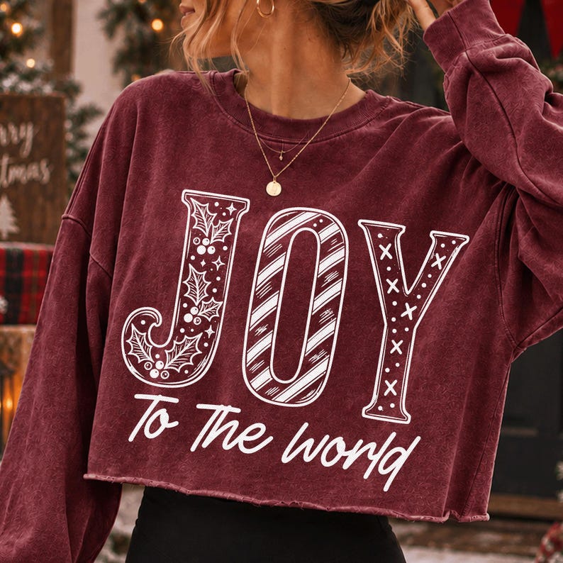 May include: A maroon long-sleeve cropped sweatshirt featuring the white text JOY, with the letter J decorated with holly leaves and berries, the O with diagonal stripes, and the Y with crosses, followed by the text To The world below.