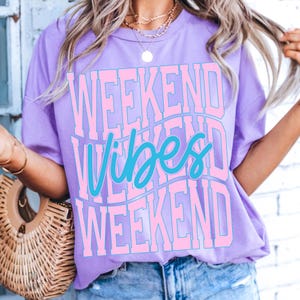 K&ouml;nnte beinhalten: Ein lilafarbenes T-Shirt mit dem Schriftzug "WEEKEND VIBES WEEKEND" in einem wellenf&ouml;rmigen Design. Der Text ist hellrosa mit dem Wort "Vibes" in t&uuml;rkisfarbener Schrift. Das Shirt hat einen Rundhalsausschnitt und kurze &Auml;rmel.