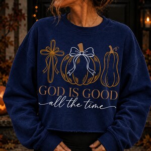 Puede incluir: Una sudadera azul marino de manga larga con un estampado gr&aacute;fico de una cruz, una calabaza con un lazo blanco y una calabaza alargada. El texto "GOD IS GOOD" aparece en negrita sobre el texto en cursiva "all the time" en color blanco.