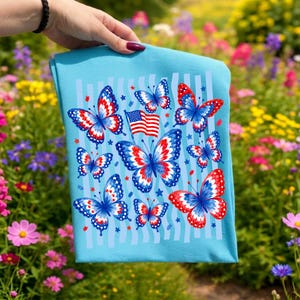 Puede incluir: Una camiseta azul claro con un estampado gr&aacute;fico de varias mariposas con patrones de alas rojos, blancos y azules. El dise&ntilde;o incluye una peque&ntilde;a bandera estadounidense y formas de estrellas dispersas sobre un fondo de rayas verticales azul claro.