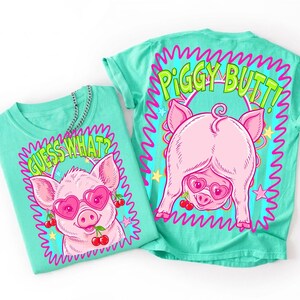 Op de afbeelding: Twee turquoise t-shirts met cartoon varkensafbeeldingen. E&eacute;n shirt heeft een varken met een hartvormige zonnebril en de tekst "GUESS WHAT?". Het andere shirt toont de achterkant van een varken met een hartvormige zonnebril en de tekst "PIGGY BUTT!".