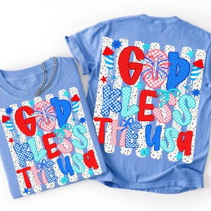 May include: A light blue short-sleeve t-shirt featuring a graphic print with the text GOD BLESS THE USA in red, white, and blue patterns. The design includes fireworks, stars, and rocket motifs over a background of vertical black and white stripes.