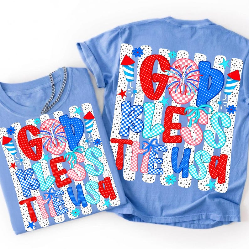 May include: A light blue short-sleeve t-shirt featuring a graphic print with the text GOD BLESS THE USA in red, white, and blue patterns. The design includes fireworks, stars, and rocket motifs over a background of vertical black and white stripes.