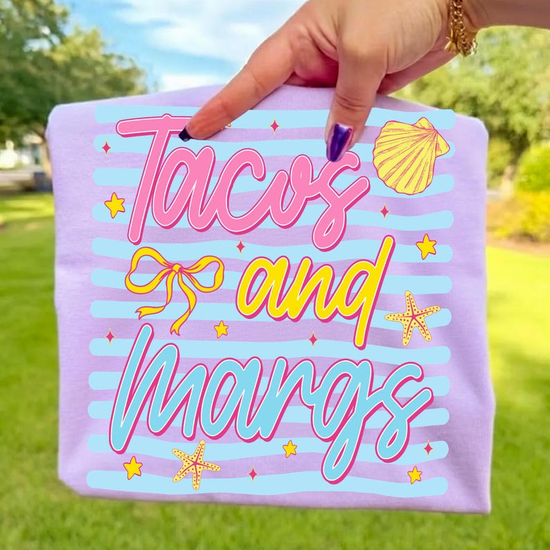 May include: A lilac t-shirt with the words "Tacos and Margs" in pink and yellow script. The shirt has blue horizontal stripes and is decorated with yellow starfish, shells, and stars. The shirt is being held up by a person.