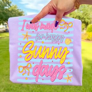 Pu&ograve; includere: Maglietta lilla con la scritta "I was made for happy sunny days" in rosa e giallo. Il design include strisce blu, stelle marine, fiocchi e una conchiglia.
