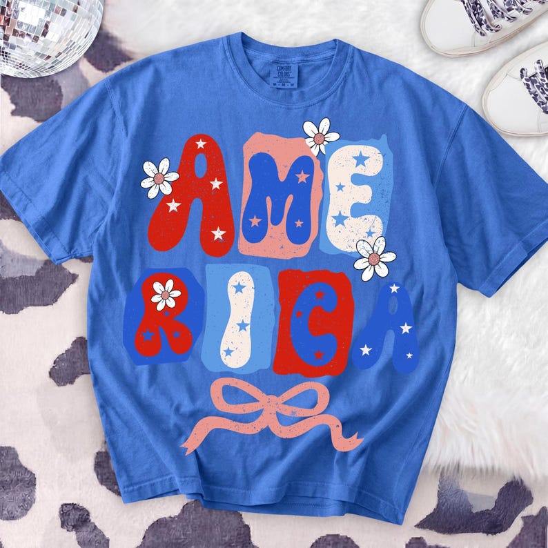 May include: A blue t-shirt with the word "AMERICA" in red, white, and blue block letters. The letters are decorated with stars and flowers. A pink ribbon is at the bottom of the design. White trainers with patterned laces are in the background.