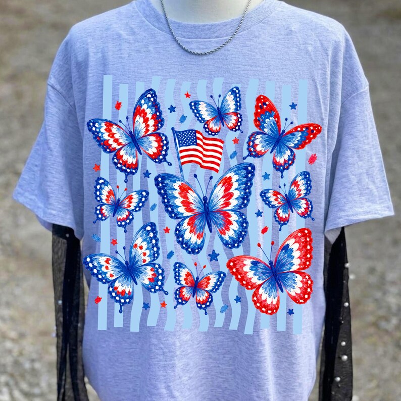 Puede incluir: Una camiseta gris claro de manga corta con un estampado gr&aacute;fico de varias mariposas en patrones rojos, blancos y azules. El dise&ntilde;o incluye una peque&ntilde;a bandera estadounidense y formas de estrellas dispersas sobre rayas verticales azul claro.