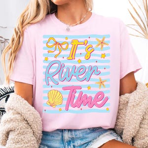 May include: Pink t-shirt with the text "It's River Time" in a colorful design. The design includes a yellow bow, starfish, a seashell, and stars. The shirt has blue horizontal stripes.