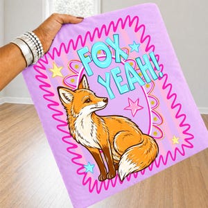 Op de afbeelding: Een rechthoekige, lichtpaarse deken met een cartoon vos illustratie. De vos is oranje en wit, met bruine poten, en de woorden "FOX YEAH!" in turquoise. Roze sterren en een roze golvende rand maken het ontwerp compleet.
