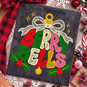 May include: A dark gray t-shirt featuring a graphic of a bell formed by the text BERRY BELLS in red, yellow, and pink, topped with a white bow and surrounded by green holly leaves and berries, with small yellow star accents.