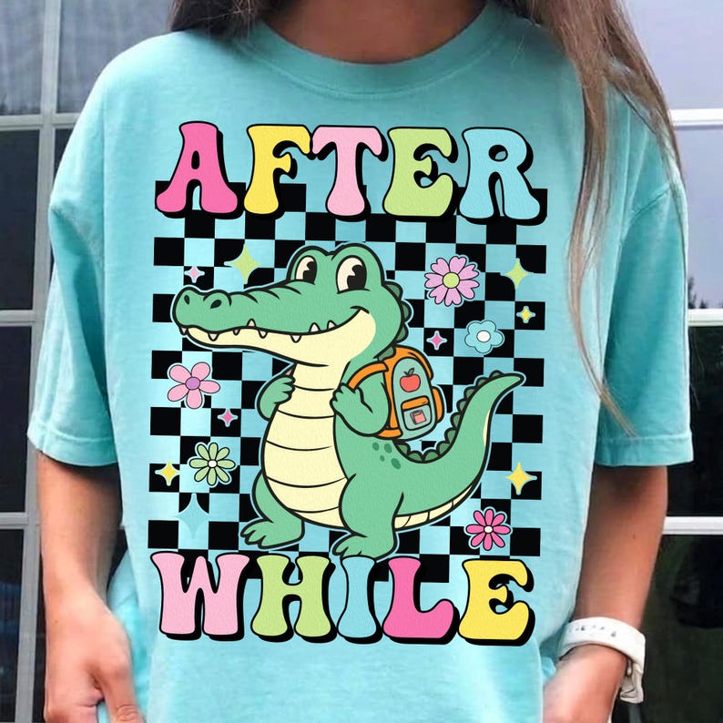 K&ouml;nnte beinhalten: Ein hellblaues T-Shirt mit den Worten "AFTER WHILE" in einer Retro-Schriftart. Das Shirt zeigt ein Cartoon-Alligator mit einem Rucksack, vor einem schwarz-wei&szlig; karierten Hintergrund mit floralen und Stern-Akzenten.