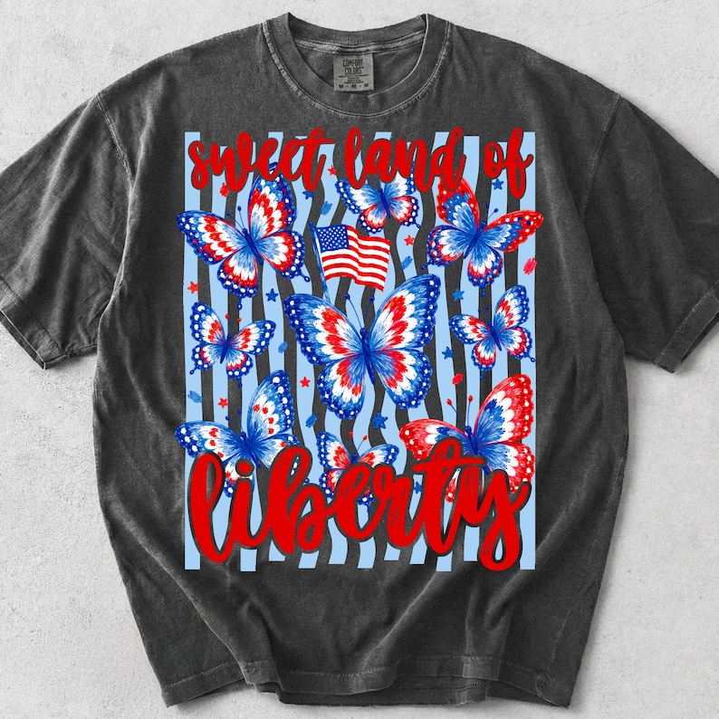 May include: A charcoal grey short-sleeve t-shirt featuring a graphic print of red, white, and blue butterflies over a light blue and black striped background. The text "sweet land of liberty" is printed in red script across the top and bottom.