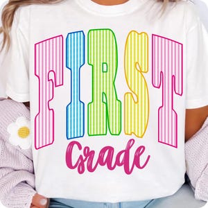 May include: A white short-sleeved t-shirt featuring the text FIRST in large, vertical-striped letters colored pink, blue, green, and yellow. Below this, the word Grade is printed in a pink cursive script font. The design is centered on the chest.