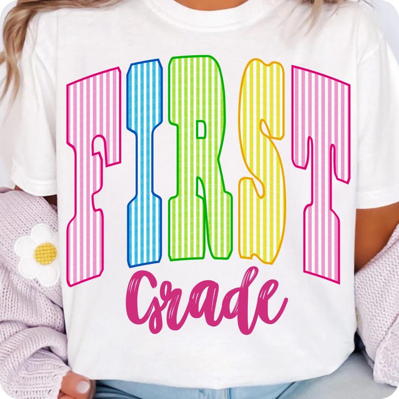 May include: A white short-sleeved t-shirt featuring the text FIRST in large, vertical-striped letters colored pink, blue, green, and yellow. Below this, the word Grade is printed in a pink cursive script font. The design is centered on the chest.